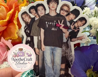 May include: A die-cut sticker featuring a group of seven individuals posing together. The central figure wears a black t-shirt and blue jeans, holding a pink microphone. The sticker includes a small circular logo with the text ApotheCraft Studio.