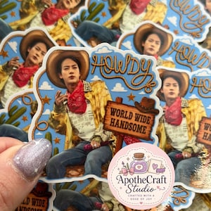 May include: A die-cut sticker featuring a person in a cowboy hat, yellow patterned shirt, and red bandana against a blue sky background with cacti. The text reads HOWDY and WORLD WIDE HANDSOME. A circular logo for ApotheCraft Studio is visible.