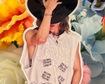 May include: A die-cut sticker featuring a person wearing a black wide-brimmed hat, a white sleeveless shirt with text and four square graphic patterns, and a black garment with white vertical stripes. The sticker has a white border edge.