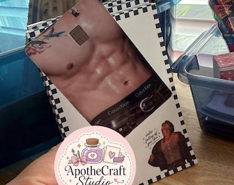 May include: A rectangular credit card skin featuring a photographic print of a muscular torso wearing Calvin Klein underwear and a belt. The design includes a black and white checkered border, a small illustration of a person, and the text water falling off your skin.
