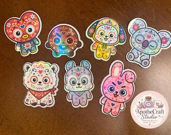 May include: A collection of seven die-cut stickers featuring cartoon characters with colorful floral patterns and a circular sticker with the text ApotheCraft Studio Crafted with a dose of joy. The characters include a heart, a round face, a dog, a koala, a bear, a rabbit, and a pink bunny.