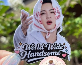 May include: A die-cut sticker featuring a person wearing a white headscarf with red patterns and a white hooded sweatshirt. The text "World Wide Handsome" is printed in black script below the person, accompanied by small pink hearts and stars.