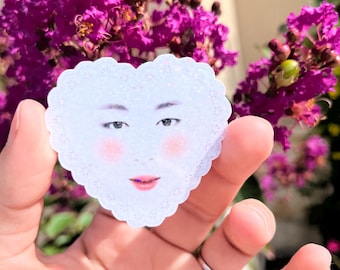 May include: A heart-shaped sticker with a scalloped lace-like edge and a holographic finish. The center features a realistic human face with dark eyebrows, almond-shaped eyes, rosy pink cheeks, and soft pink lips, all printed on a white background.