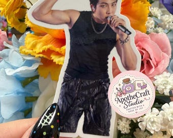 May include: A die-cut sticker featuring a person with dark hair wearing a black sleeveless top and dark pants, holding a microphone while flexing one arm. A circular pink logo with the text ApotheCraft Studio Crafted with a Dose of Joy is attached.