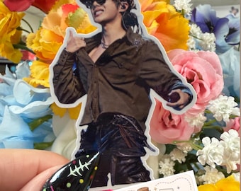 May include: A die-cut sticker featuring a person wearing a brown button-down shirt, dark pants, a leopard print cap, and sunglasses. The sticker is positioned in front of a colorful floral arrangement and a small rectangular label reading ApotheCraft Studio.