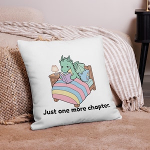Op de afbeelding: Wit vierkant kussen met een cartoon draak die in bed leest. De draak is groen met blauwe vleugels, onder een gestreepte deken. De tekst "Just one more chapter" staat eronder.