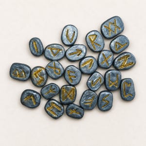 May include: A collection of twenty-four small, smooth, dark gray stones, each featuring a unique, hand-painted gold rune symbol. The stones are oval or rectangular in shape with rounded edges, arranged in a scattered cluster on a neutral surface.