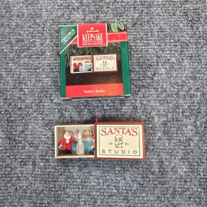 May include: A Hallmark Keepsake Ornament titled Santa's Studio, featuring a miniature matchbox-style box with a sliding tray. The box displays the text SANTA'S STUDIO 1991. Inside, a small Santa figure stands next to a white figure in a workshop.