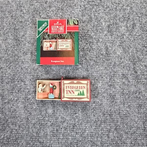 May include: A Hallmark Keepsake Ornament titled Evergreen Inn 1991, featuring a miniature matchbox-style box. The box displays a scene of a person at a front desk and a guest, with the text Evergreen Inn 1991 printed on the sliding cover.
