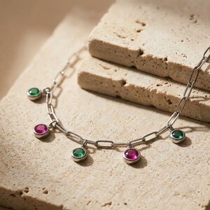 Op de afbeelding: Een zilverkleurige ketting met een reeks afwisselend ronde, in zettingen geplaatste edelstenen in levendige tinten smaragdgroen en fuchsia roze. De ketting is opgebouwd uit langwerpige, rechthoekige schakels voor een gebalanceerd ontwerp.