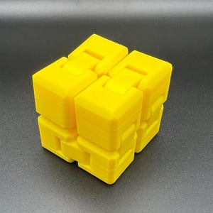 May include: A bright yellow fidget cube, a puzzle toy, is displayed against a dark background. The cube is composed of several connected blocks that can be manipulated and rotated. The toy is designed to provide a tactile and engaging experience.