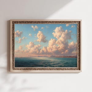 Könnte beinhalten: Gerahmtes Landschaftsgemälde, das eine ruhige Meereslandschaft unter einem blauen Himmel mit flauschigen, weißen und rosa Wolken zeigt. Das Meer ist tief türkis und der Rahmen hellbeige mit aufwendigen Details.