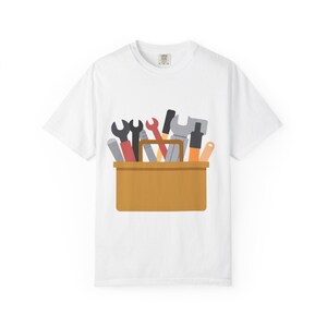 Könnte beinhalten: Weißes T-Shirt mit einer Cartoon-Werkzeugkiste. Die braune Werkzeugkiste enthält verschiedene Werkzeuge, darunter Schraubenschlüssel, einen Hammer und Schraubendreher in Grau, Schwarz, Rot und Orange. Das T-Shirt hat einen Rundhalsausschnitt.