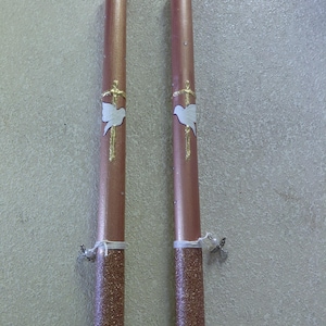 May include: Two tall, cylindrical rose gold candles featuring a gold painted cross and a white dove emblem. The base of each candle is covered in rose gold glitter, and the top is accented with a gold beaded trim. A small silver angel charm is tied to each.