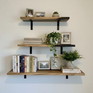 Puede incluir: Tres estantes de madera fijados a una pared blanca, soportados por soportes metálicos negros. Los estantes exhiben libros, fotos enmarcadas y plantas en macetas, creando una exhibición decorativa. Los estantes son de madera natural.