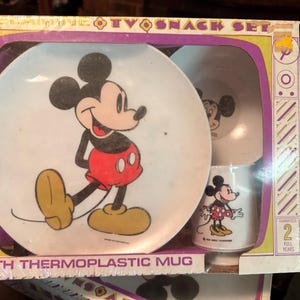 May include: Vintage Mickey Mouse TV snack set in original packaging. The set includes a plate, a bowl, and a mug, all featuring Mickey Mouse. The box has the text "MEGAVISION TV SNACK SET" and "WITH THERMOPLASTIC MUG".