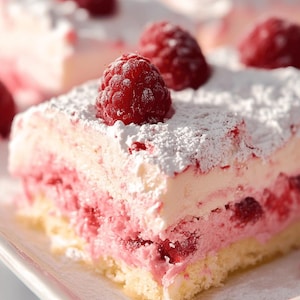 May include: A square slice of layered raspberry cake featuring a light sponge base, a thick pink raspberry cream filling with visible fruit pieces, a smooth white cream topping, and a dusting of icing sugar, garnished with fresh raspberries.