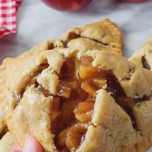 May include: A golden-brown baked apple hand pie with a crimped edge, broken open to reveal a warm, glistening filling of diced apples coated in a dark cinnamon glaze, with two whole red apples visible in the blurred background.