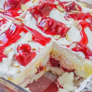 May include: A square dessert cake in a glass dish featuring layers of white sponge, red fruit filling, and a thick topping of white whipped cream. The top is decorated with swirls of bright red cherry glaze and scattered sliced almond pieces.
