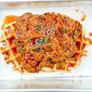 Può includere: Un petto di pollo fritto dorato posto sopra una cialda quadrata, entrambi conditi con una glassa rosso-arancio. Il pollo è guarnito con erba cipollina verde e semi di sesamo bianchi, il tutto servito in un vassoio rettangolare di metallo.