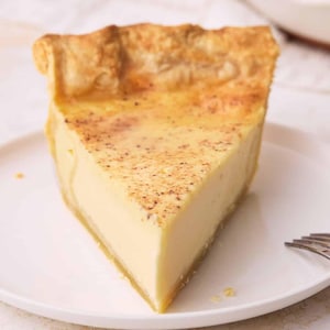 May include: A single triangular slice of custard pie on a white plate. The pie features a smooth, pale yellow custard filling with a light dusting of brown spice on top, encased in a golden-brown, flaky, crimped pastry crust edge.