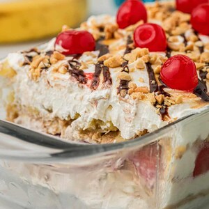 Może przedstawiać: Deser typu banana split w prostokątnym szklanym naczyniu, zwieńczony białą bitą śmietaną, jasnoczerwonymi wiśniami maraschino, posiekanymi orzeszkami ziemnymi i polewą z ciemnej czekolady, z widocznymi warstwami owoców i kremu wewnątrz naczynia.
