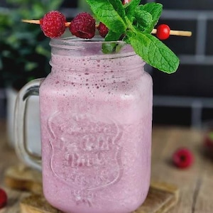 May include: A refreshing pink smoothie in a mason jar, garnished with fresh raspberries, mint leaves, and a red and white striped straw. The drink sits on a wooden surface, with additional raspberries scattered around.