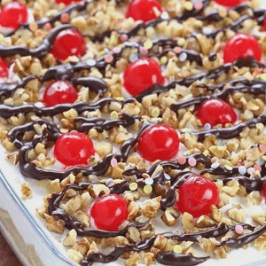 May include: A rectangular glass dish containing a layered dessert topped with white frosting, chopped walnuts, bright red maraschino cherries, drizzled dark chocolate sauce, and small multicolored round sprinkles scattered across the surface.