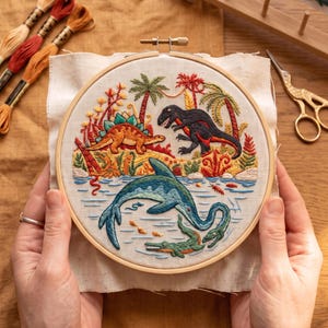 Könnte beinhalten: Ein runder Stickrahmen aus Holz mit einem Stoffeinsatz, der eine detaillierte, handgestickte prähistorische Szene zeigt. Das Motiv umfasst einen braunen Stegosaurus, einen schwarzen Tyrannosaurus, einen blauen Plesiosaurus und ein grünes Krokodil.