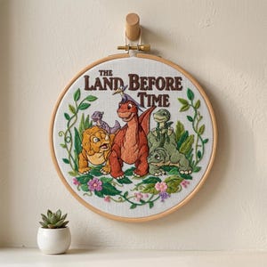 May include: Embroidered hoop art featuring characters from "The Land Before Time." The design includes several dinosaurs in shades of orange, yellow, and green, surrounded by green vines and pink flowers. The title of the movie is embroidered in brown letters.