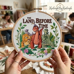 May include: Embroidered hoop art featuring characters from "The Land Before Time" with the text of the movie title. The artwork showcases a vibrant scene with dinosaurs, flowers, and green foliage, all set against a white fabric background within a wooden hoop.