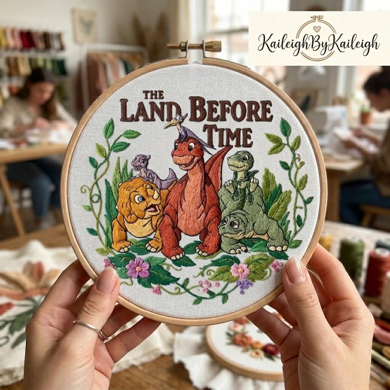 May include: Embroidered hoop art featuring characters from "The Land Before Time" with the text of the movie title. The artwork showcases a vibrant scene with dinosaurs, flowers, and green foliage, all set against a white fabric background within a wooden hoop.