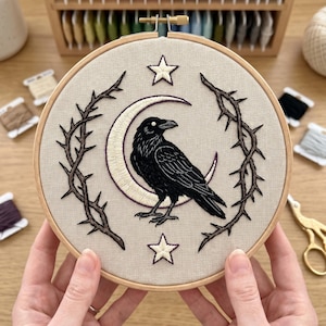 May include: A circular wooden embroidery hoop containing a beige fabric panel with a hand-stitched design of a black raven perched in front of a cream-colored crescent moon, framed by two brown thorny branches and two white stars with purple outlines.