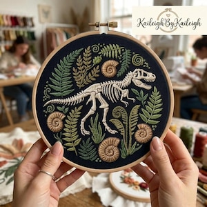 Peut inclure: Broderie sur tambour représentant un squelette de Tyrannosaurus Rex entouré de fougères vertes et de motifs de fossiles sur fond bleu marine. L'œuvre est tenue par des mains. Le texte "Kaileigh By Kaileigh" est visible.