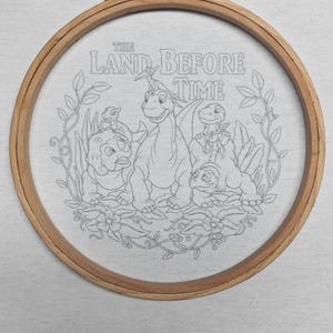 May include: Embroidery hoop featuring a line drawing of characters from "The Land Before Time." The design includes dinosaurs and the text "THE LAND BEFORE TIME." The wooden hoop is secured with a brass clasp, set against a light gray fabric.