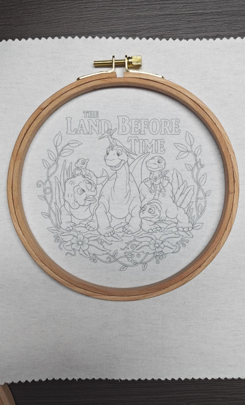 May include: Embroidery hoop featuring a line drawing of characters from "The Land Before Time." The design includes dinosaurs and the text "THE LAND BEFORE TIME." The wooden hoop is secured with a brass clasp, set against a light gray fabric.