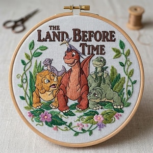 May include: Embroidered hoop art featuring characters from "The Land Before Time." The design includes several dinosaurs in shades of orange, yellow, and green, surrounded by green foliage and pink flowers. The title is stitched in brown.