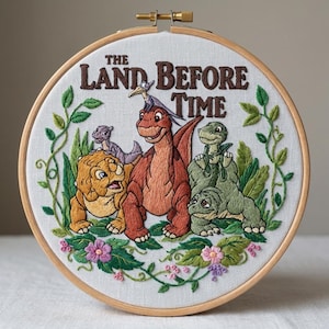 May include: Embroidered hoop art featuring characters from "The Land Before Time." The design includes several dinosaurs in shades of orange, green, and yellow, surrounded by embroidered foliage and flowers. The title is stitched in brown thread.