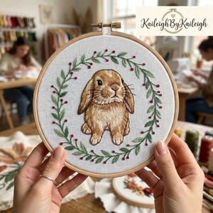 May include: Embroidered bunny hoop art. A brown bunny is centered on a white fabric, surrounded by a green and red floral wreath. The embroidery is set in a wooden hoop. The brand name "Kaileigh By Kaileigh" is in the top right corner.