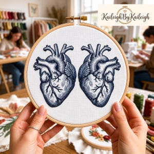 Op de afbeelding: Twee borduurringen met anatomische harten, geborduurd met blauw garen op witte stof. De ringen zijn van hout. De tekst "Kaileigh By Kaileigh" is zichtbaar. Andere borduurprojecten zijn op de achtergrond te zien.