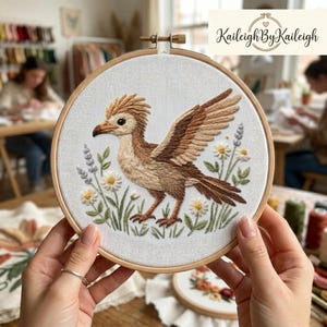 May include: Embroidered hoop art featuring a detailed bird with outstretched wings, in shades of brown and tan, surrounded by embroidered daisies and lavender. The artwork is set against a white fabric background, held in a wooden embroidery hoop. The text "Kaileigh By Kaileigh" is in the upper right corner.