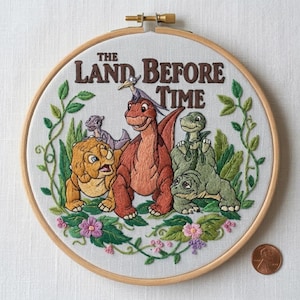 May include: Embroidered hoop art featuring characters from "The Land Before Time." The design includes several dinosaurs in shades of orange, yellow, and green, surrounded by green foliage and pink flowers. The title is embroidered in brown.