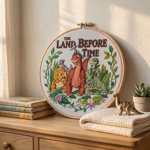May include: Embroidered hoop art featuring characters from "The Land Before Time" with the title in brown text. The design includes a red dinosaur, green dinosaurs, and other characters surrounded by green leaves and pink flowers.