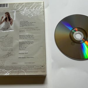 K&ouml;nnte beinhalten: Eine Delta Goodrem DVD-H&uuml;lle und eine Disc. Die R&uuml;ckseite zeigt ein Foto der K&uuml;nstlerin, eine Titelliste mit Live at VJ HQ, Video Clips, Behind the Scenes, Growing Up und Extras sowie eine gedruckte Notiz und eine Unterschrift. Die Disc ist rund.