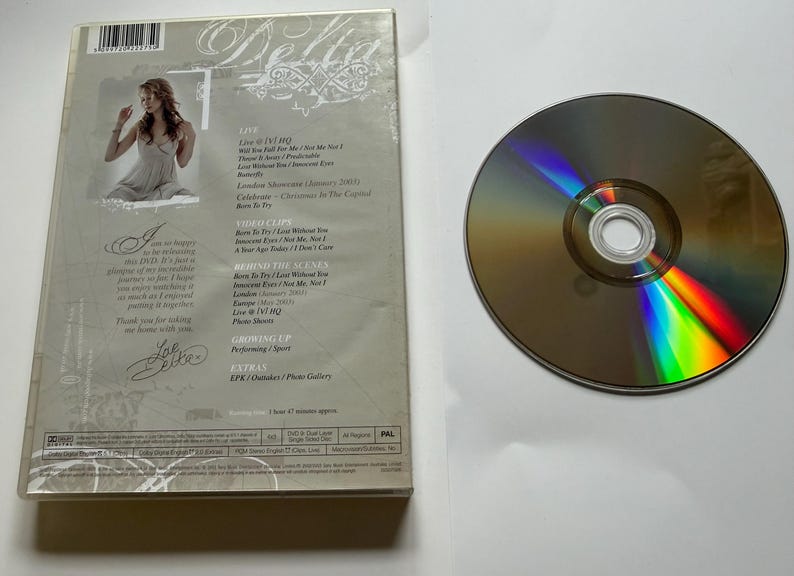 K&ouml;nnte beinhalten: Eine Delta Goodrem DVD-H&uuml;lle und eine Disc. Die R&uuml;ckseite zeigt ein Foto der K&uuml;nstlerin, eine Titelliste mit Live at VJ HQ, Video Clips, Behind the Scenes, Growing Up und Extras sowie eine gedruckte Notiz und eine Unterschrift. Die Disc ist rund.