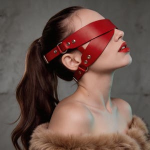 Op de afbeelding: Een rode leren blinddoek met verstelbare banden voorzien van zilverkleurige metalen gespen, klinknagels en ringen. Het ontwerp bestaat uit twee kruisende banden die om het hoofd lopen, vastgezet met een riem achter de oren.
