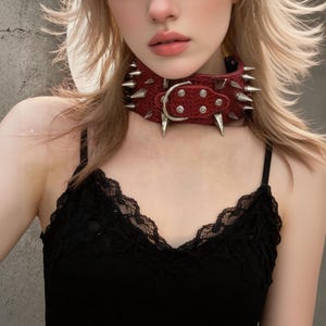 May include: A red leather choker with silver spikes and a buckle. The choker has a textured surface and is secured with a silver buckle. The spikes are evenly spaced around the collar.