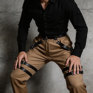 May include: Black leather leg harness with adjustable buckles and a belt with silver rings. The harness is worn over tan pants and paired with a black button-down shirt. The outfit is set against a gray concrete wall.