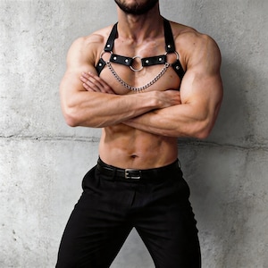 May include: Black leather body harness with silver chain and ring details. The harness features adjustable straps and a central ring with a chain. The design is a bold accessory.