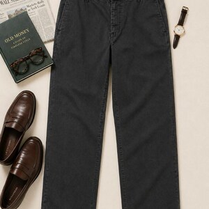 May include: Dark grey trousers with a button closure, alongside a watch, brown leather loafers, a newspaper, a book titled "OLD MONEY", and spectacles. The trousers are the central element of the image.